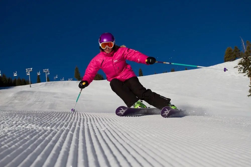 Skier in pink jacket and purple helmet skiing on groomed snow under a clear blue sky.